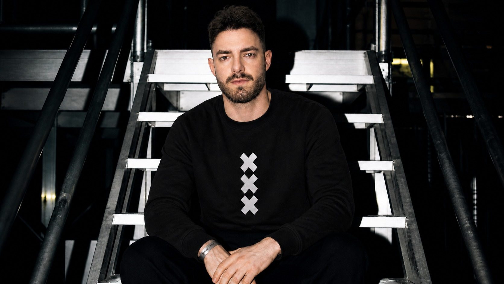 Chris Bonet seated on a metal staircase, wearing a black sweater with three white crosses